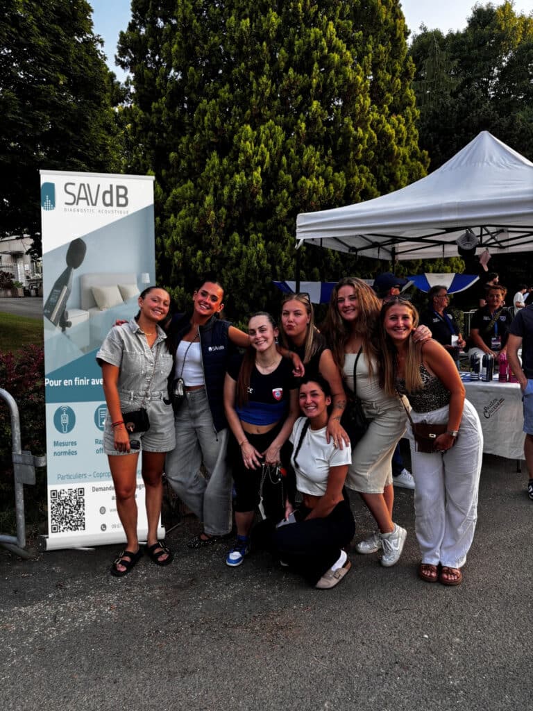 Photo de groupe prise en extérieur lors d’un événement. Plusieurs participantes posent ensemble devant un roll-up SAVdB de diagnostic acoustique installé près d’un stand.